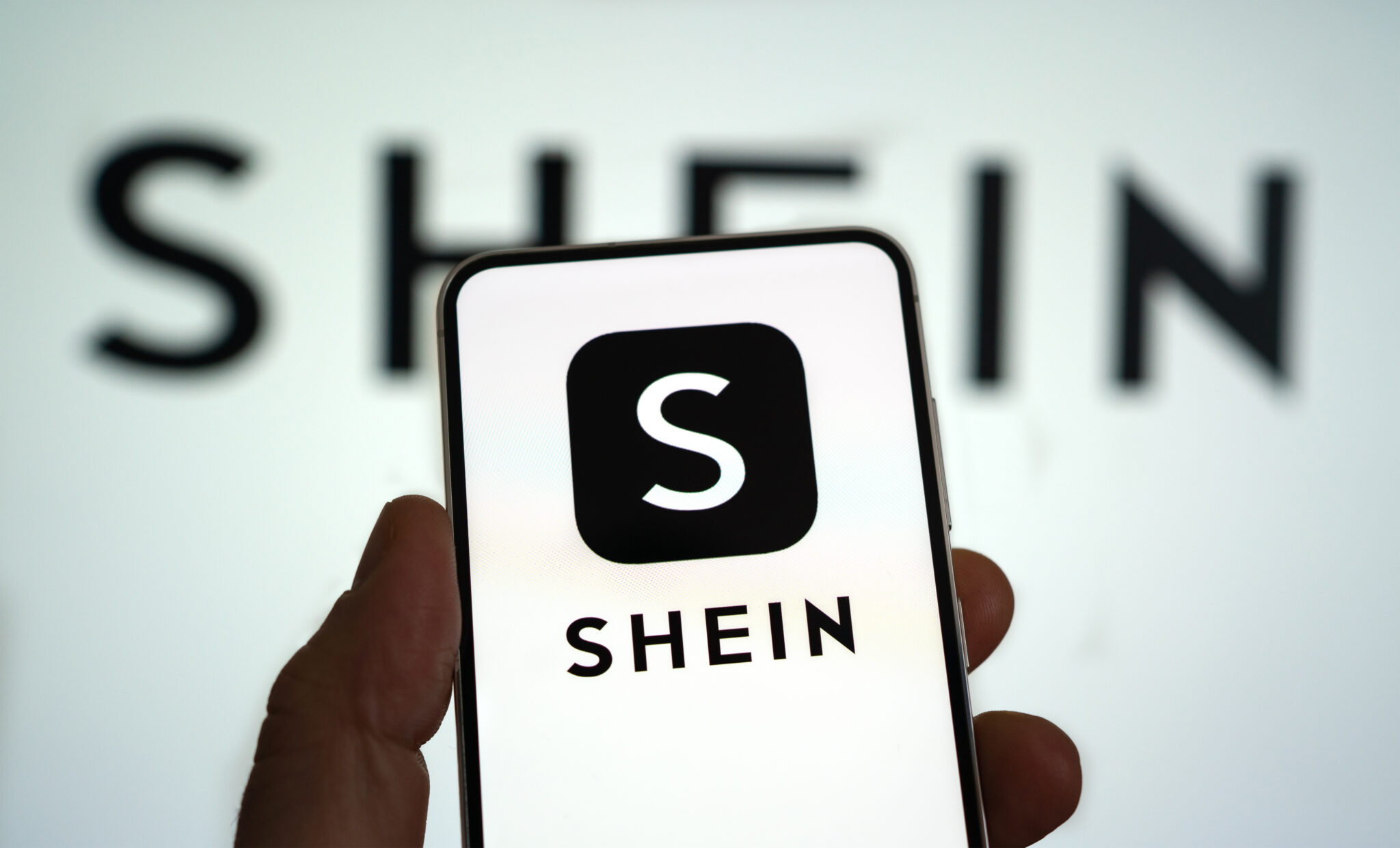 Logo marki Shein na ekranie smartfona, trzymanym na tle rozmytego logotypu marki.