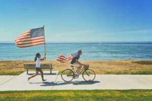 Mężczyzna na rowerze z flagą USA i dziewczynka biegnąca z drugą flagą na tle oceanu