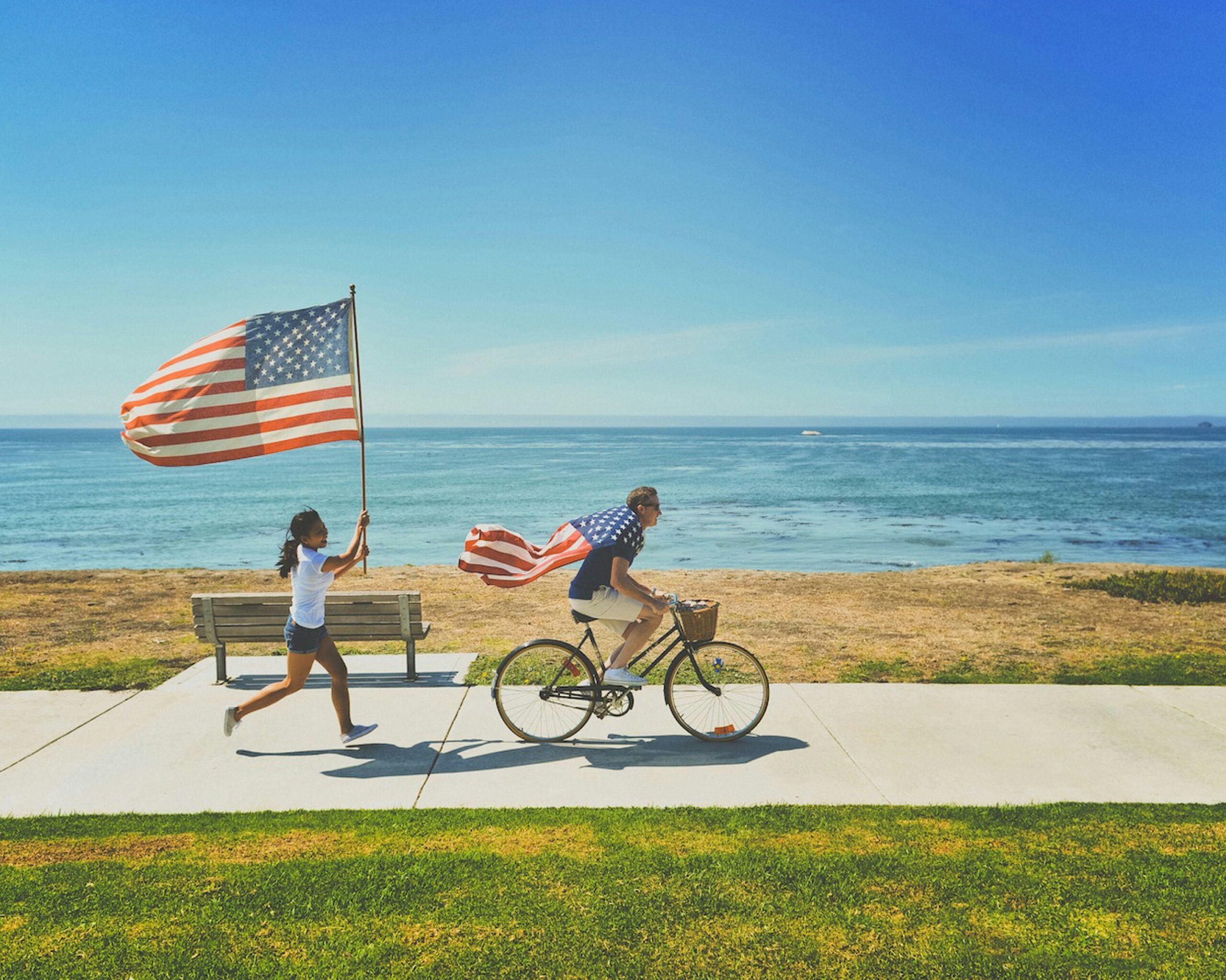 Mężczyzna na rowerze z flagą USA i dziewczynka biegnąca z drugą flagą na tle oceanu