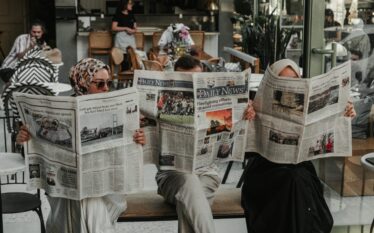 Trzy osoby siedzące w kawiarni i czytające gazety, symbolizujące rolę mediów w walce z dezinformacją klimatyczną