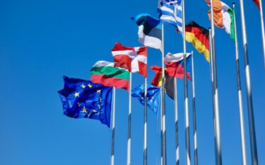 Flagi państw Unii Europejskiej oraz flaga UE na maszach na tle czystego nieba.