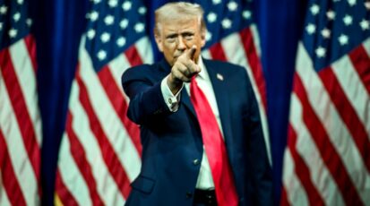 Donald Trump wskazuje palcem podczas wystąpienia na tle flag USA