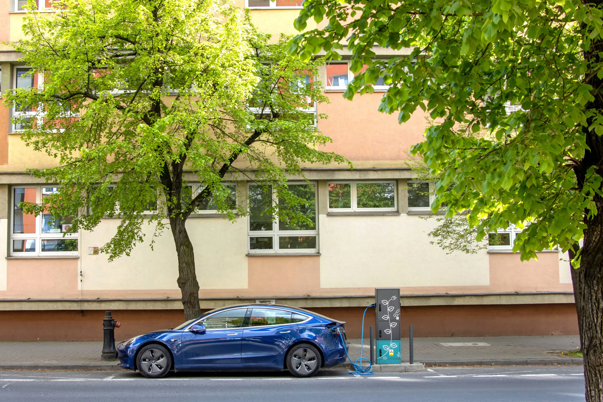 Samochód elektryczny podłączony do ładowarki przy ulicy w polskim mieście, w tle budynek mieszkalny i zielone drzewa.